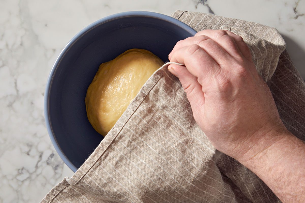overhead shot of a hand gently pulling back a striped cloth to reveal a round ball of dough in a deep blue bowl, placed on a light colored marble surface