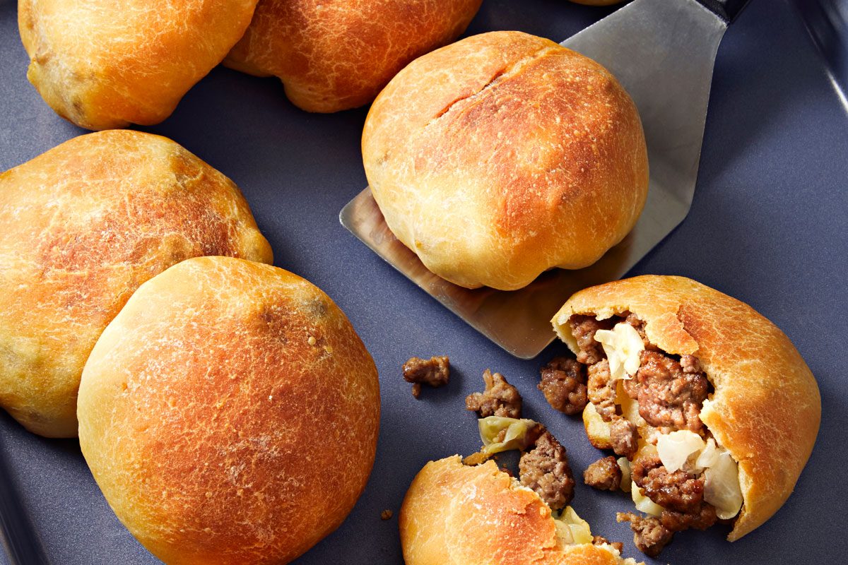closeup shot of a platter of meat buns; one of the buns has been cut open, revealing filling of ground meat and cabbage; a spatula is placed beside the opened bun