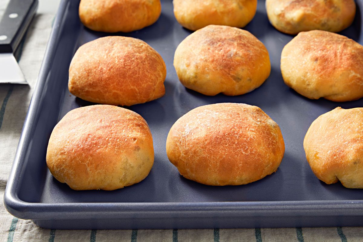 3/4th shot of a tray filled with freshly baked meat buns; they are arranged neatly on a dark blue baking tray, the background includes a kitchen towel