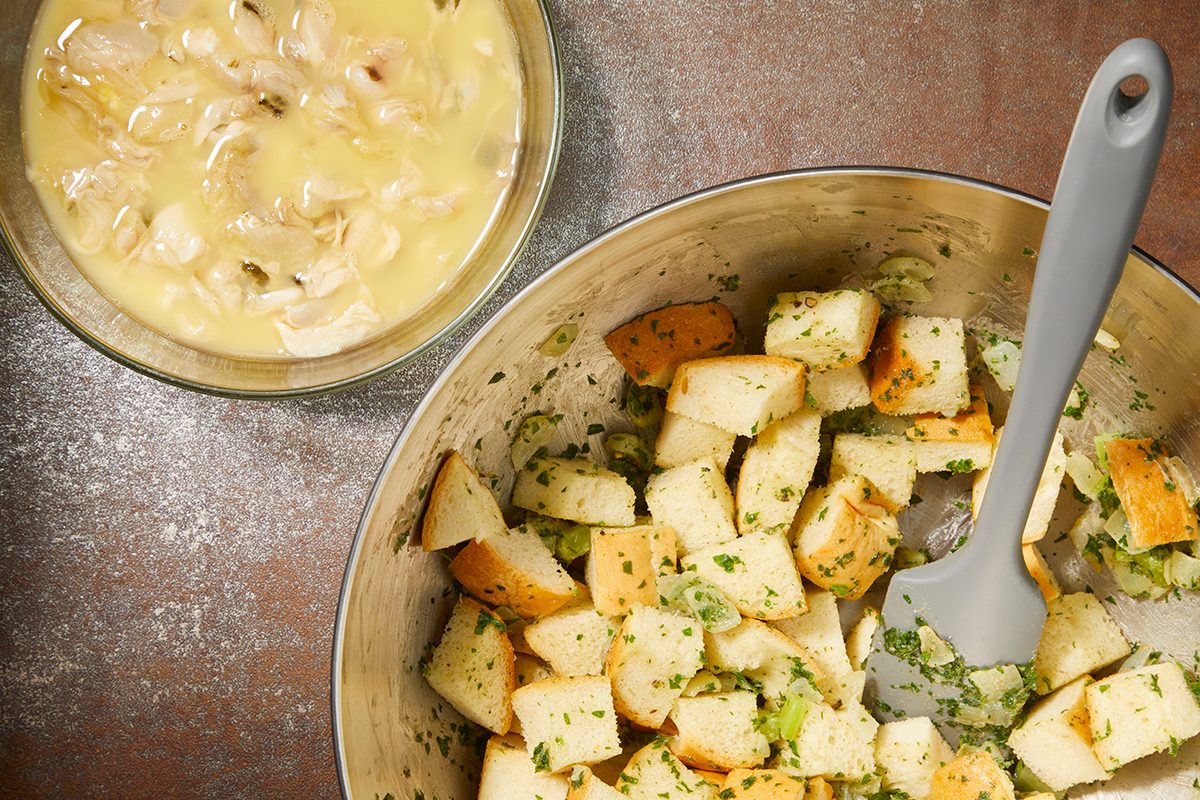 A stainless steel bowl contains cubed bread mixed with herbs and celery. A light grey spatula rests inside the bowl. Next to it, a clear bowl holds a mixture of chicken pieces and cream sauce. Both are on a brown surface.
