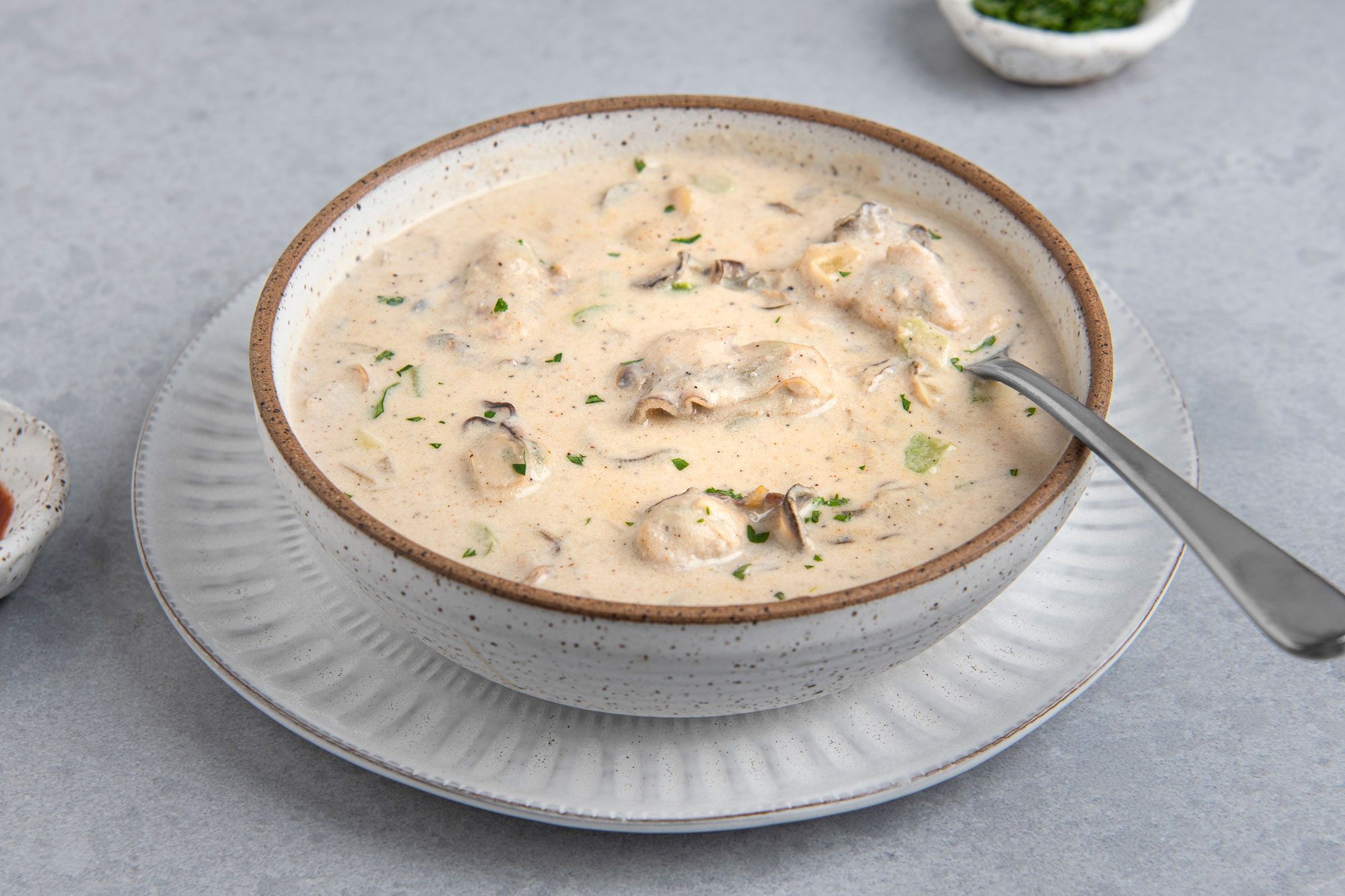 3/4th shot of a bowl of Oyster Stew on a plate with a spoon in the bowl; the bowl is a speckled white with a brown rim; the stew is a creamy white with some green bits of parsley and some dark bits of pepper