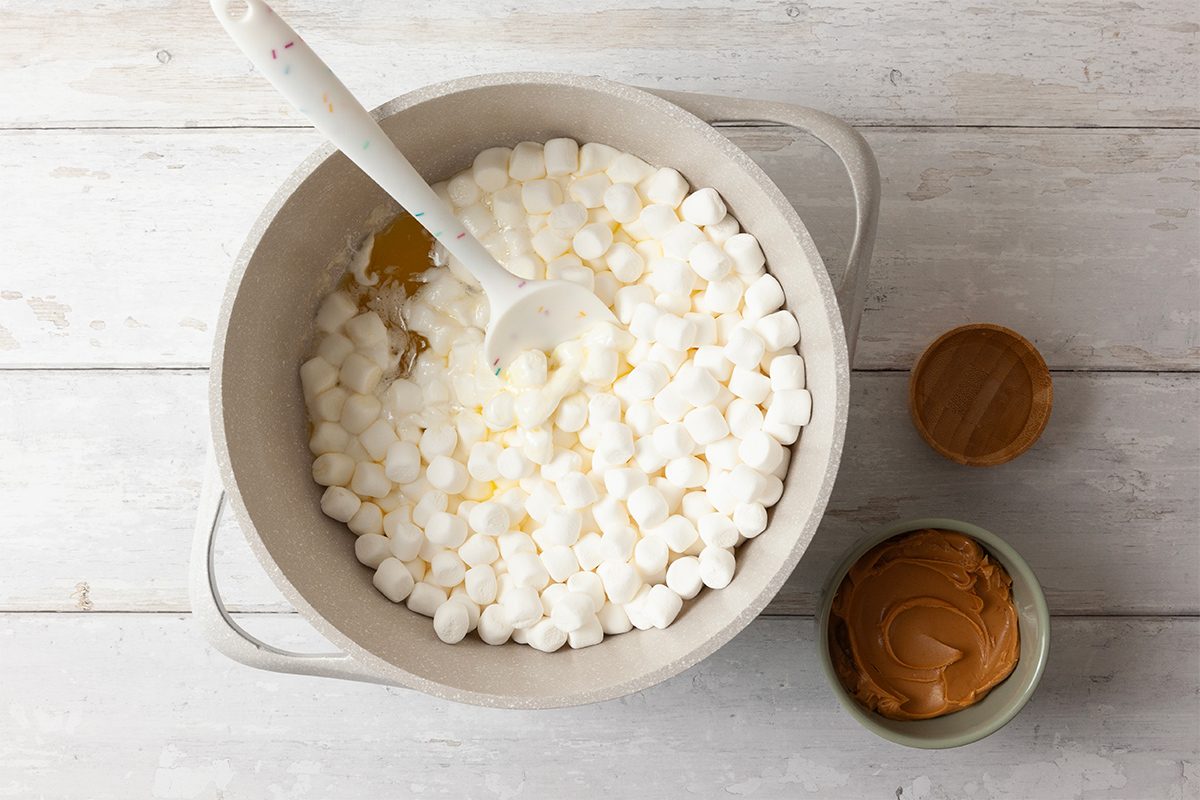 A large bowl filled with mini marshmallows and a spoon on a white wooden surface, with a small bowl of peanut butter next to it.
