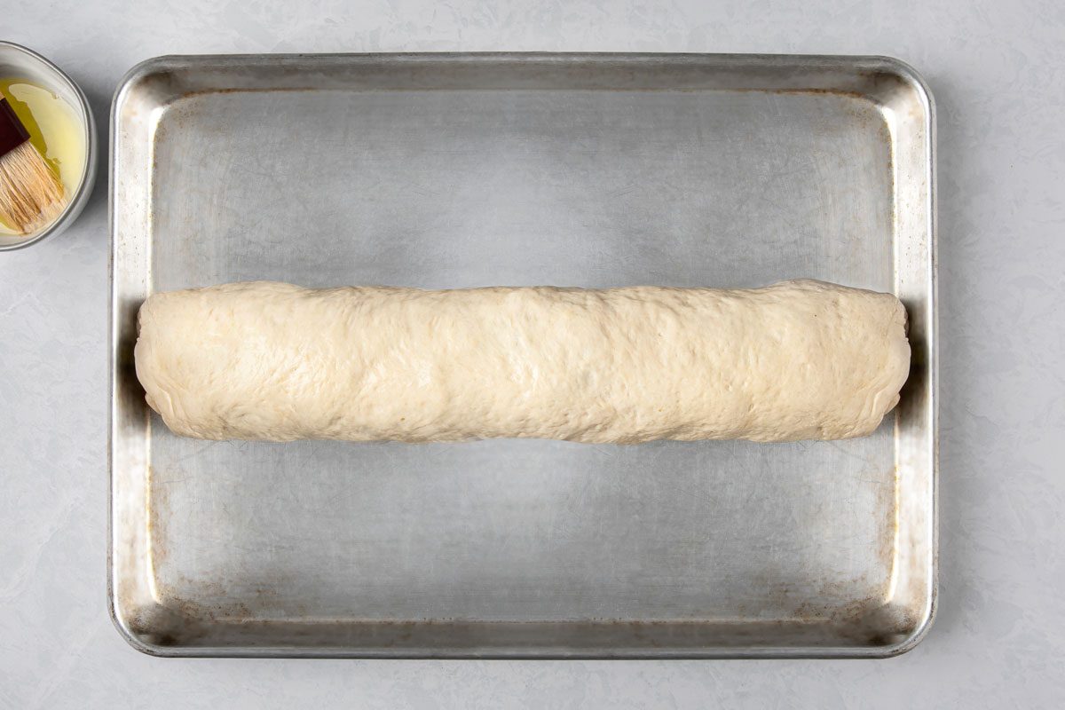 overhead shot of a rolled up a rectangular pizza on a baking tray, the baking tray is metallic and sits on a plain grey background;