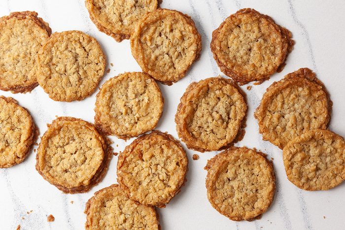 A scattering of round, golden-brown cookies on a white marble surface. The cookies have a textured appearance, likely oatmeal or similar ingredients. The arrangement is informal and slightly overlapping.