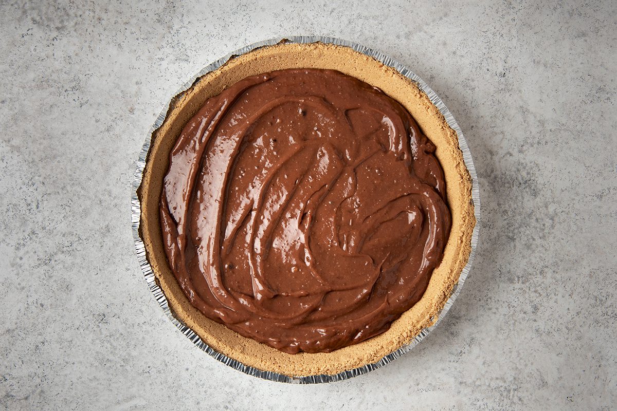 A chocolate cream pie with a smooth, rich filling sits in a metal pie tin. The crust is light brown and evenly browned, contrasting with the dark, glossy chocolate filling. The surface of the pie is textured with swirls.