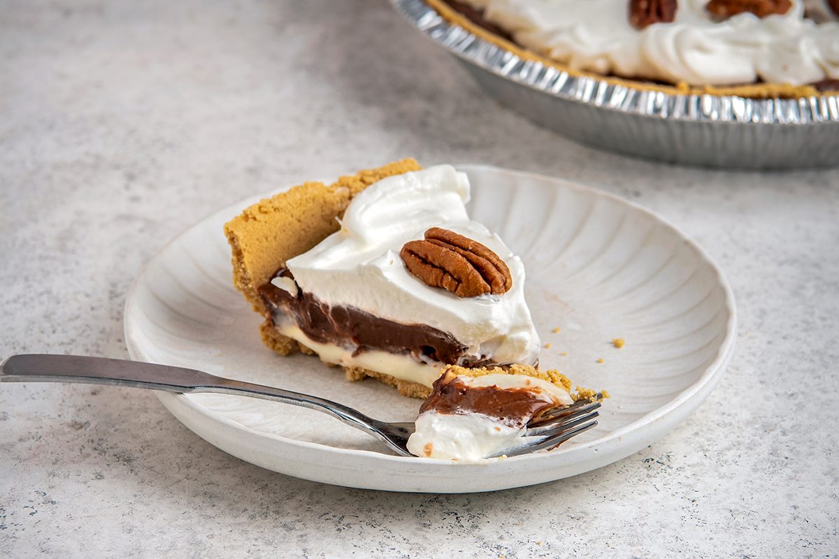 A slice of cream pie with a graham cracker crust and a chocolate filling, topped with whipped cream and a pecan. The pie slice is on a white plate with a fork, and the remaining pie is in the background.
