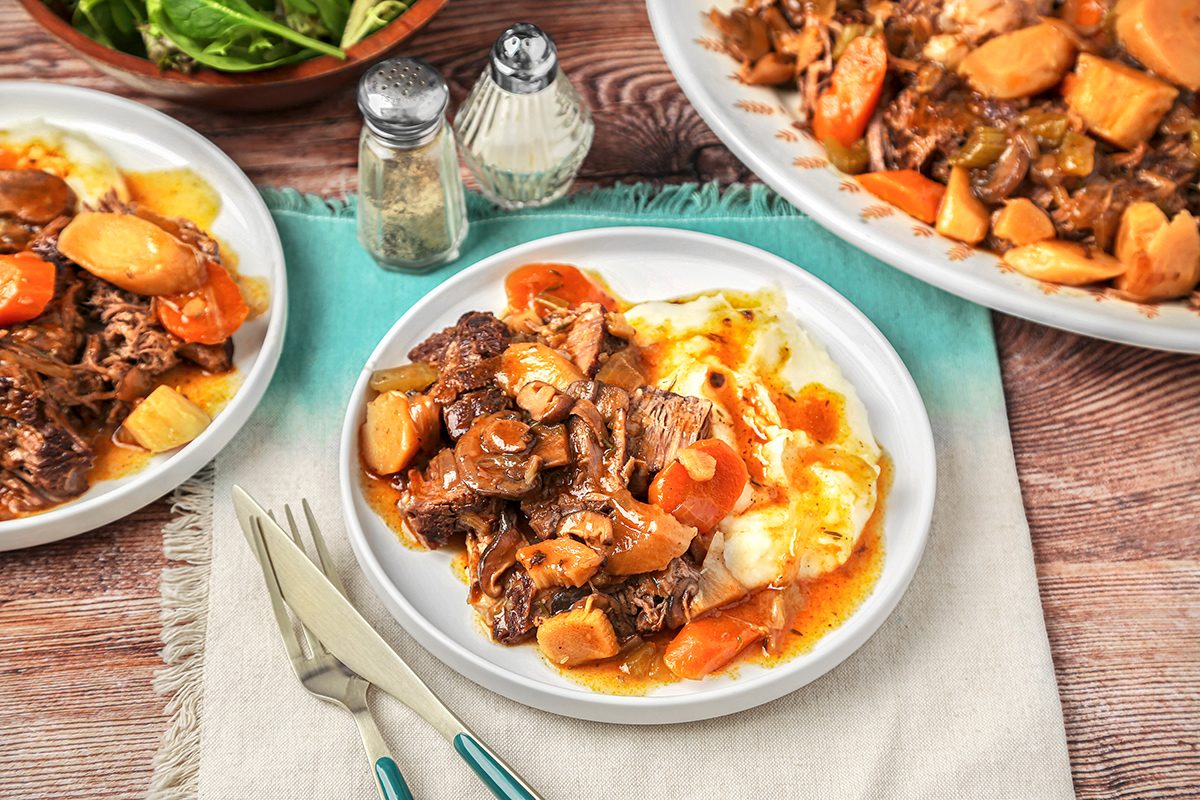 Plate of beef stew with carrots, potatoes, and gravy served over mashed potatoes. Two plates and a bowl of salad are in the background. Salt and pepper shakers, a knife, and a fork are placed next to the main plate on a napkin.