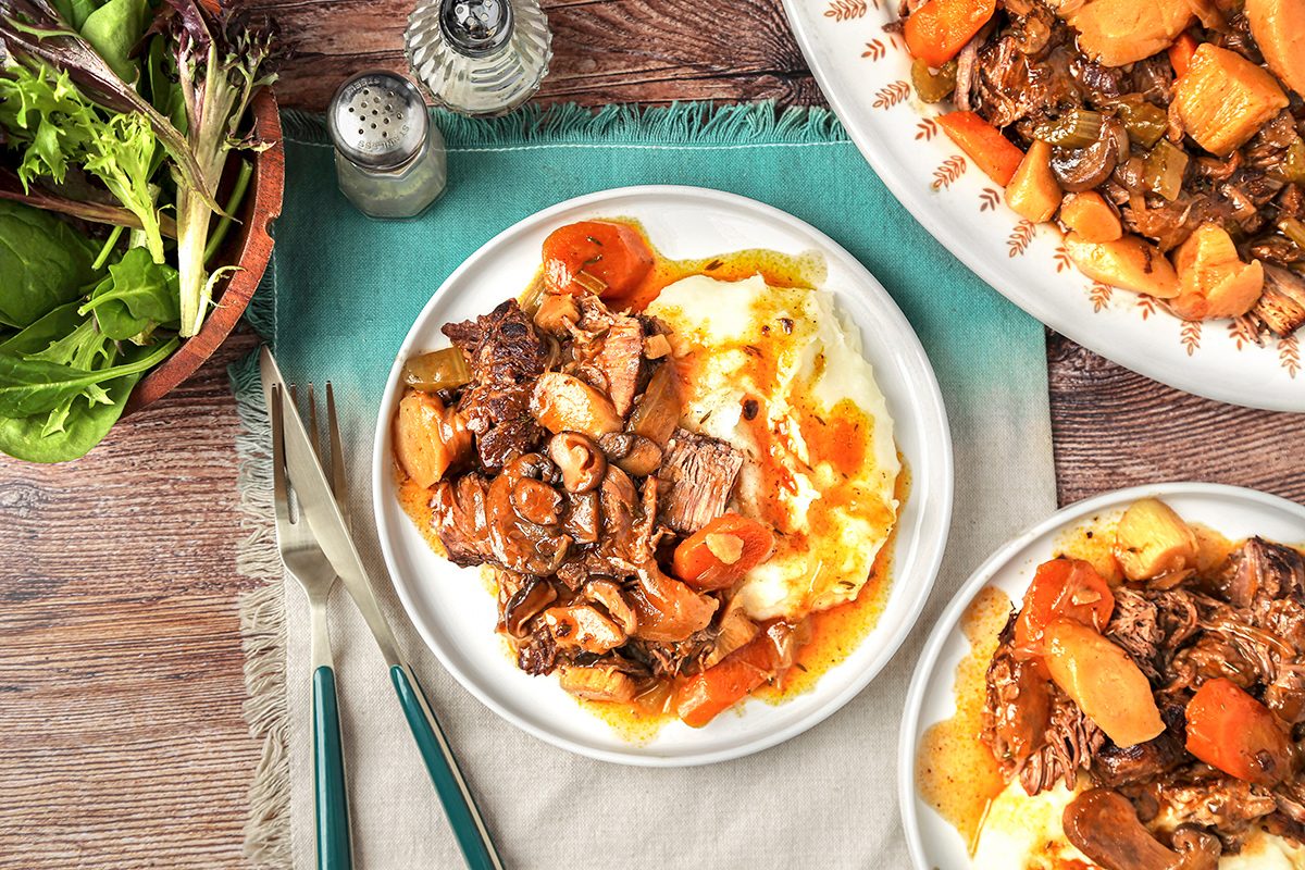 A table setting with plates of beef stew over mashed potatoes, surrounded by a bowl of fresh greens. The stew contains chunks of beef, carrots, and other vegetables in a rich sauce. Salt and pepper shakers and utensils are nearby.