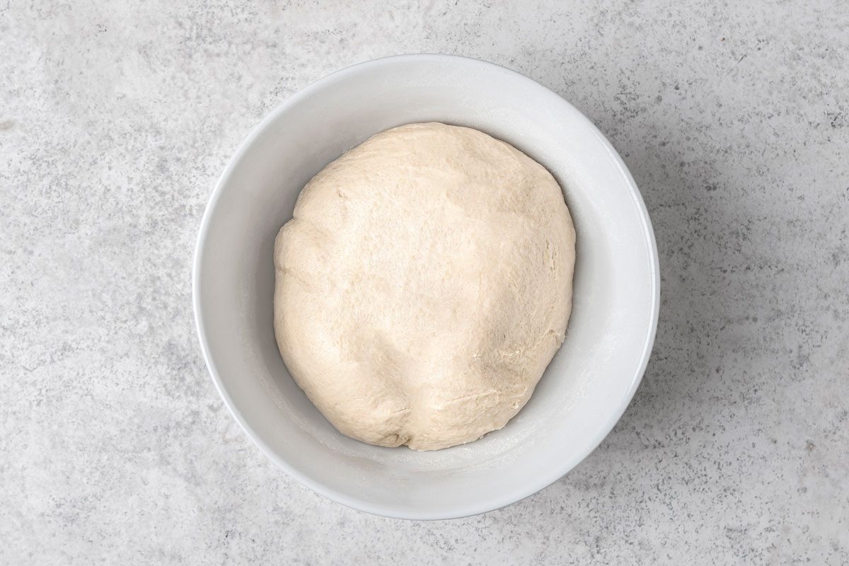 overhead shot of a ball of dough resting in a white oval shaped bowl, and is placed on a mottled grey surface