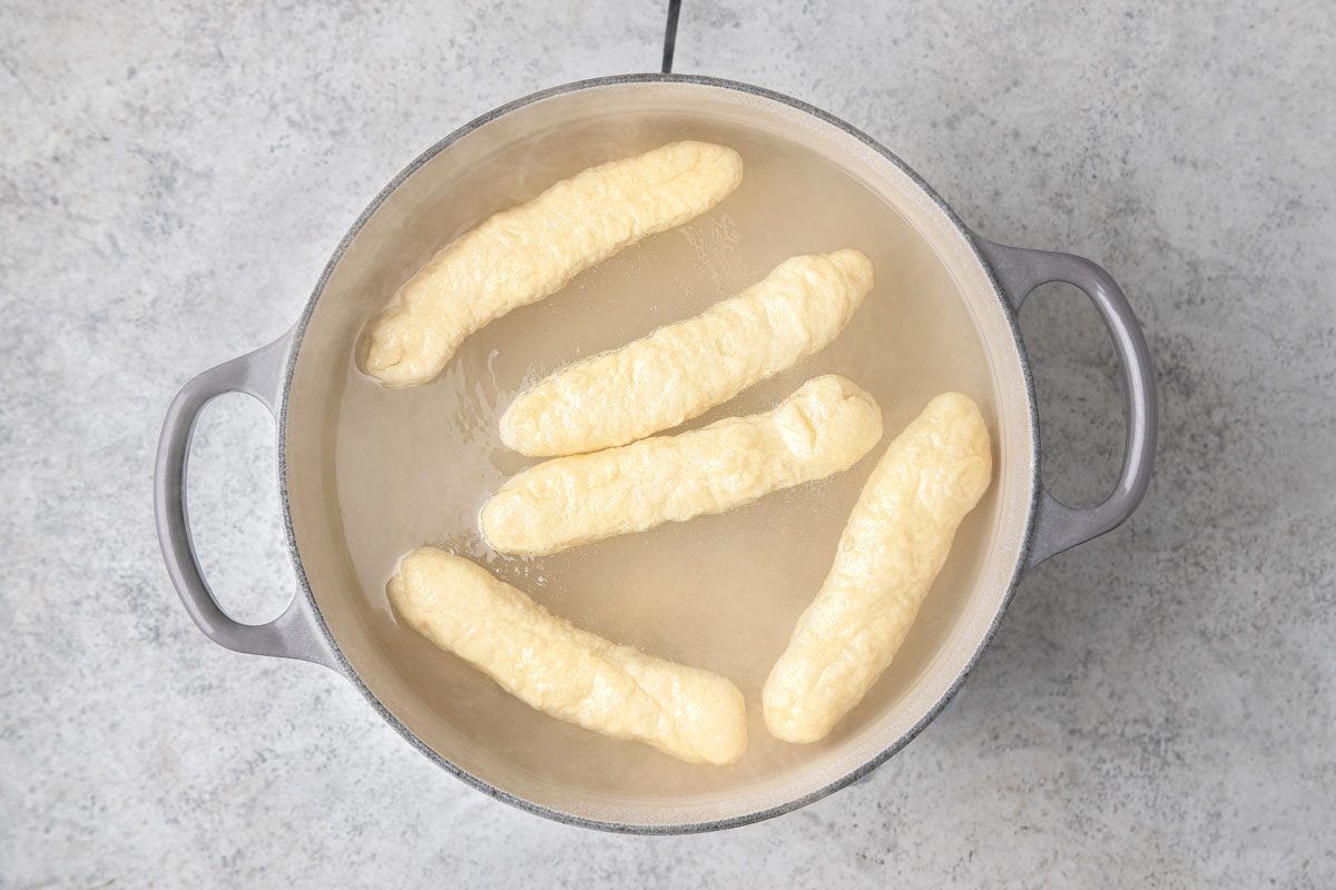 overhead shot of several oblong pale yellow pretzel sticks cooking in a pot of simmering water, the pretzel sticks are submerged in the water, the pot is grey with two handles and is placed on a grey countertop surface;