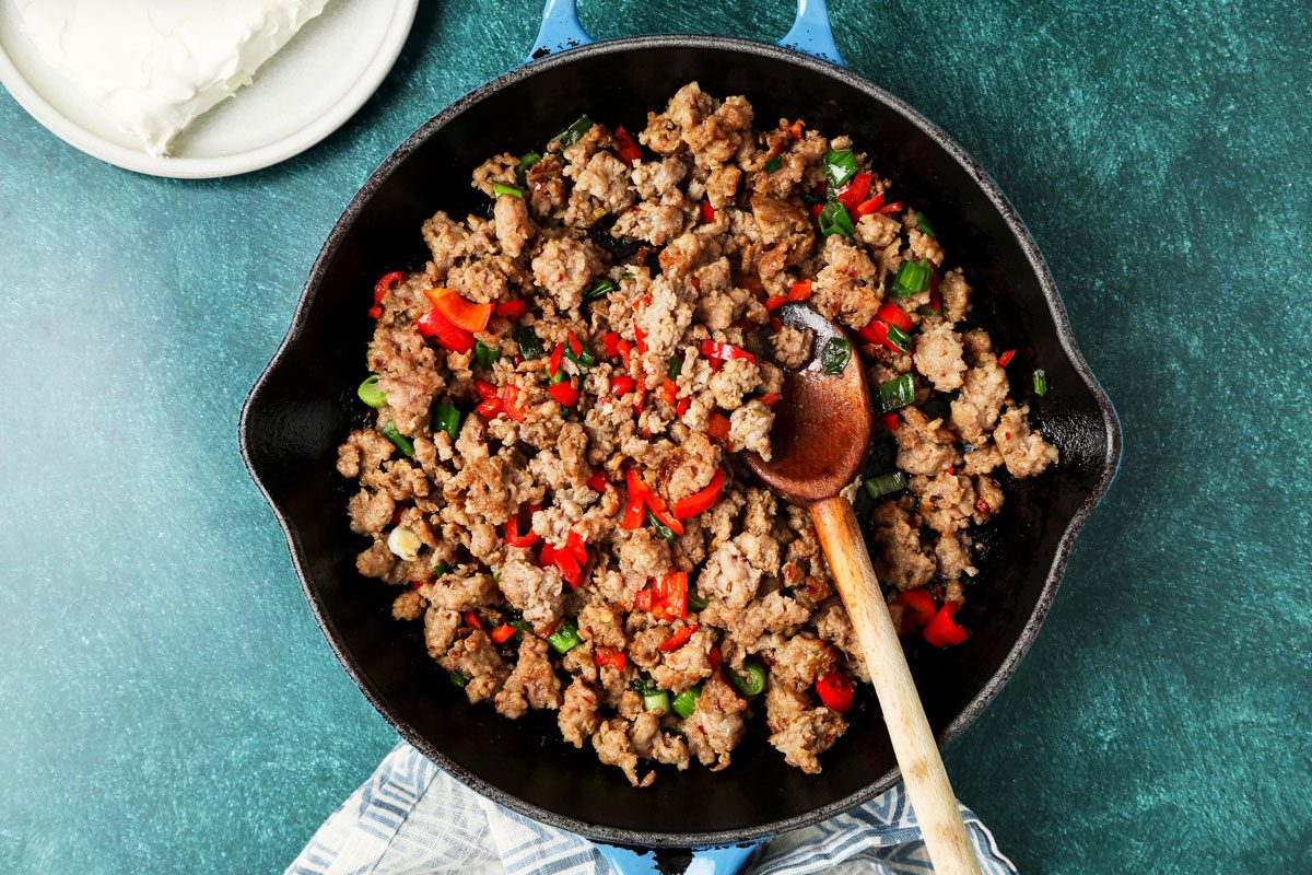 overhead shot of a cast iron skillet filled with cooked sausage, red peppers, and green onions; a wooden spoon sits inside the skillet, resting on the side; the skillet is set on a teal surface, and a white dish with a block of cream cheese sits off to the left