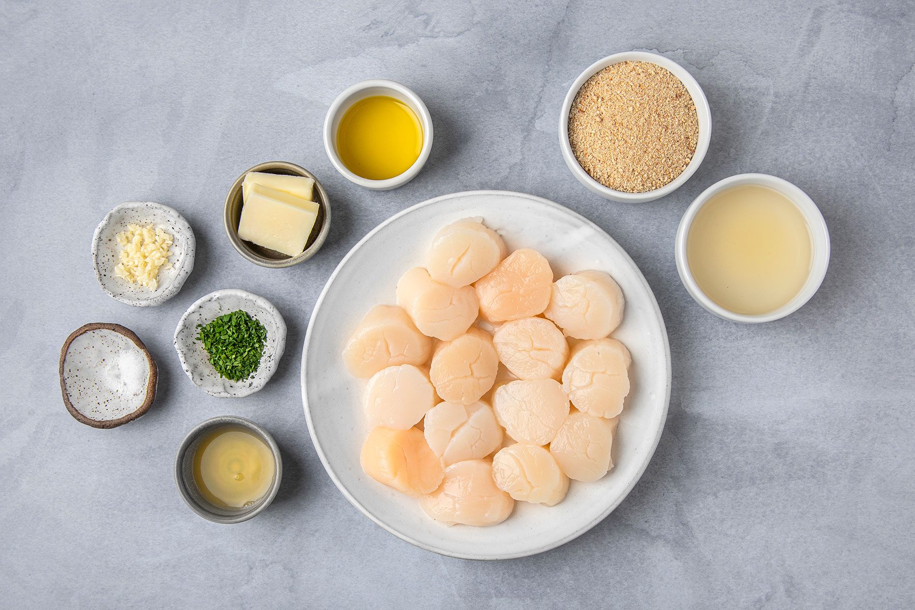 Seared Scallops ingredients olive oil, bread crumbs, white wine, butter, minced garlic, chopped herbs, lemon juice, and salt, all placed on a light gray surface.