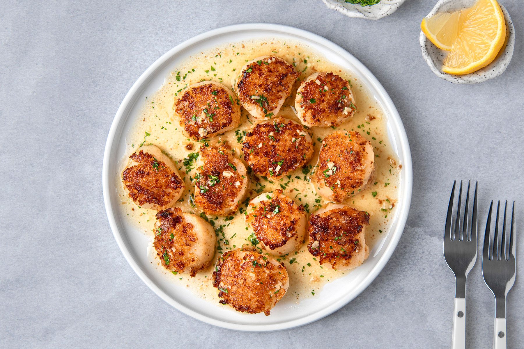 Plate of seared scallops garnished with chopped herbs, served in a creamy sauce. A lemon wedge is placed nearby, along with two forks.