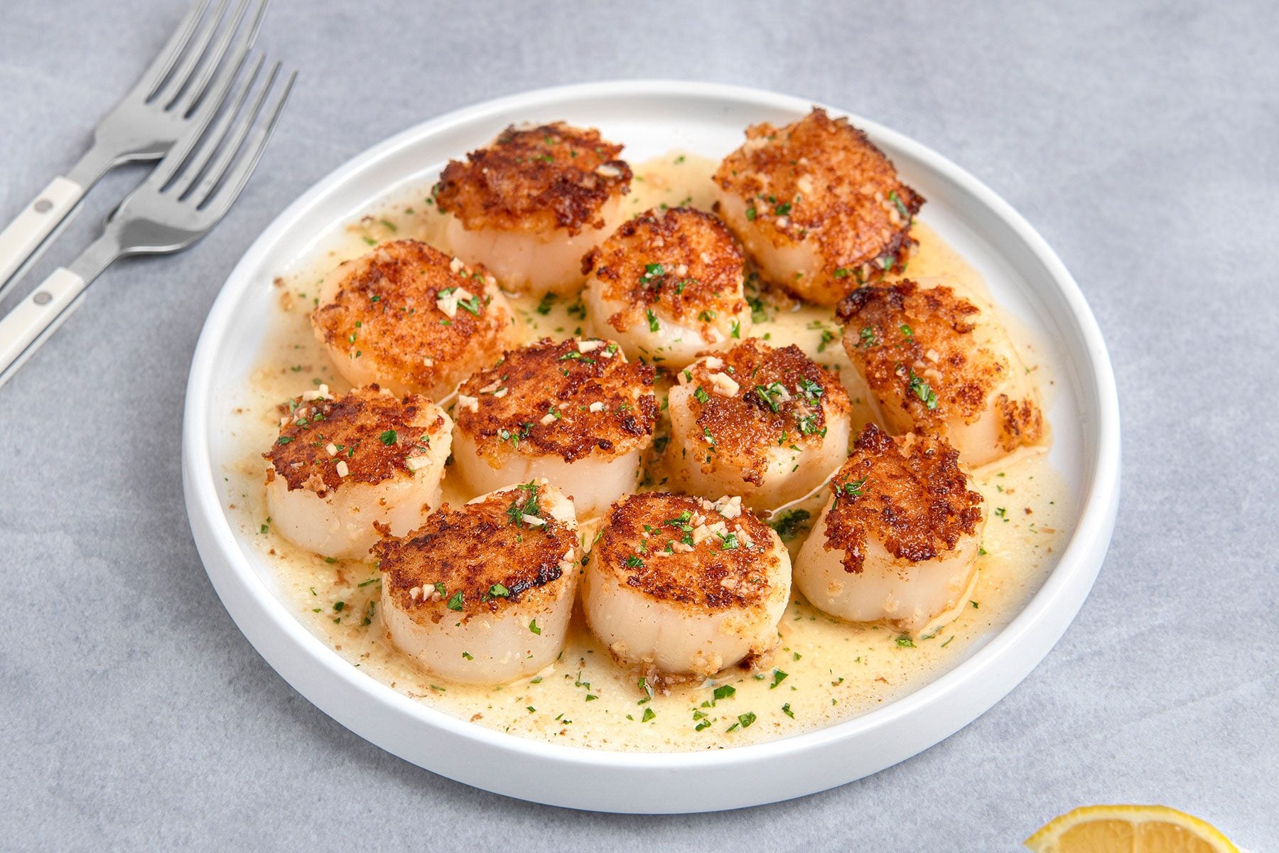A white plate with seared scallops garnished with chopped herbs and a light sauce. Two forks rest nearby on a gray surface, and a slice of lemon is partially visible in the corner.