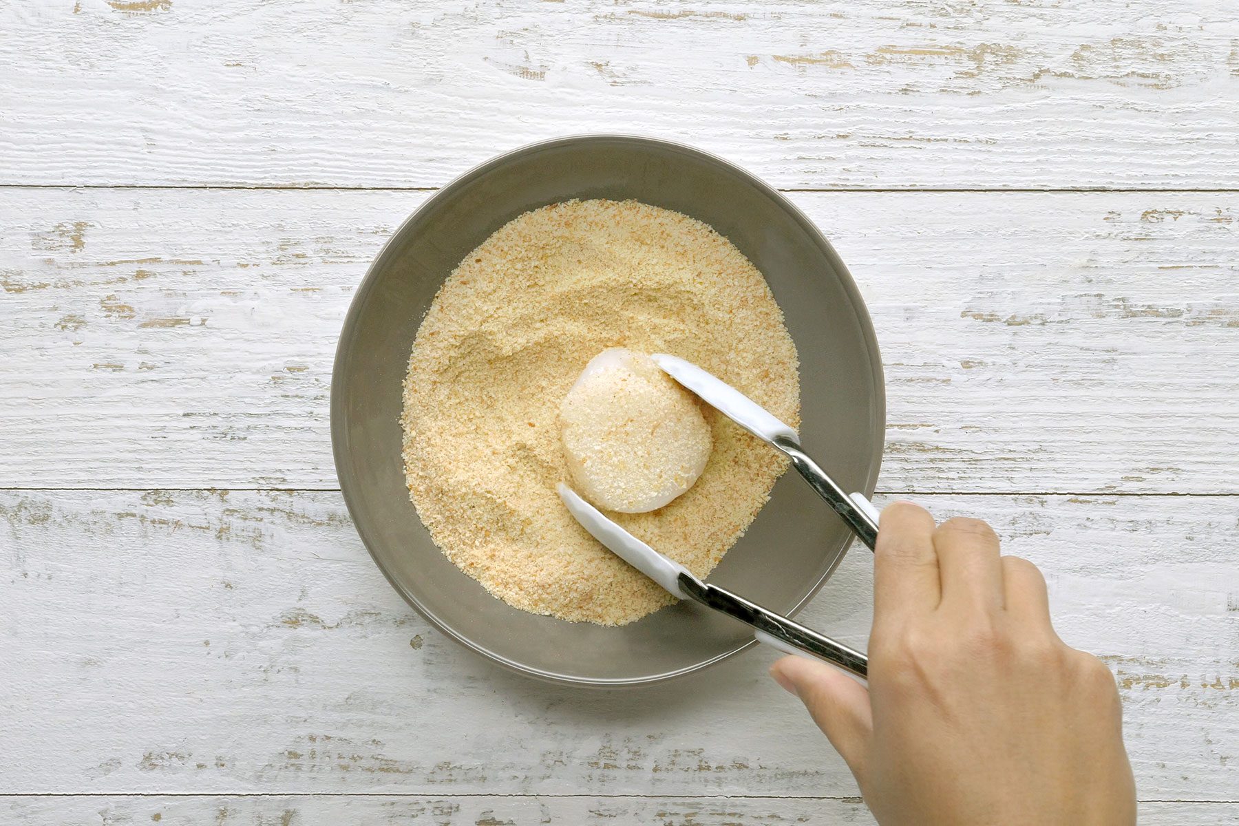 A hand uses tongs to coat a round piece of food in breadcrumbs inside a gray bowl. The bowl is set on a light wooden surface.