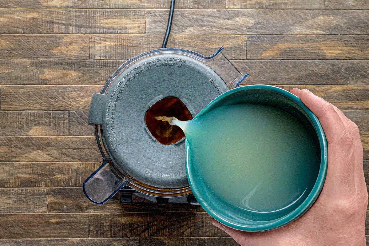 overhead shot of a blender with a green bowl being poured into it; the blender lid is on; the blender is on a wooden surface