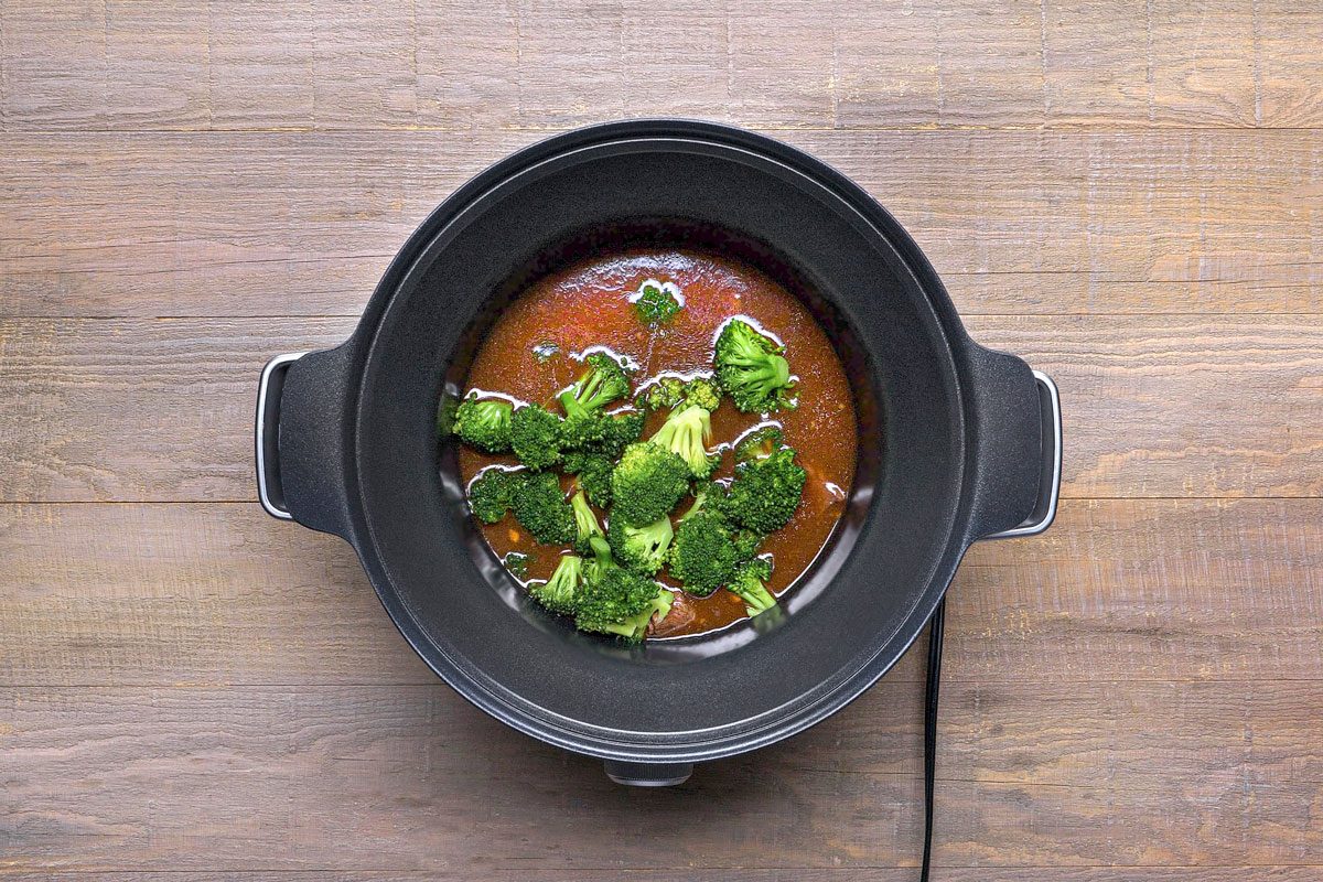 overhead shot of a black slow cooker filled with a brown liquid and broccoli florets; the slow cooker is sitting on a wooden table