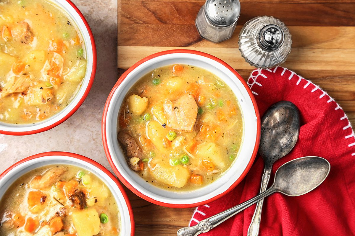 Three bowls of hearty soup with chicken, potatoes, carrots, and peas sit on a wooden surface. A red cloth napkin, two vintage spoons, and salt and pepper shakers are nearby. The bowls are white with red rims.