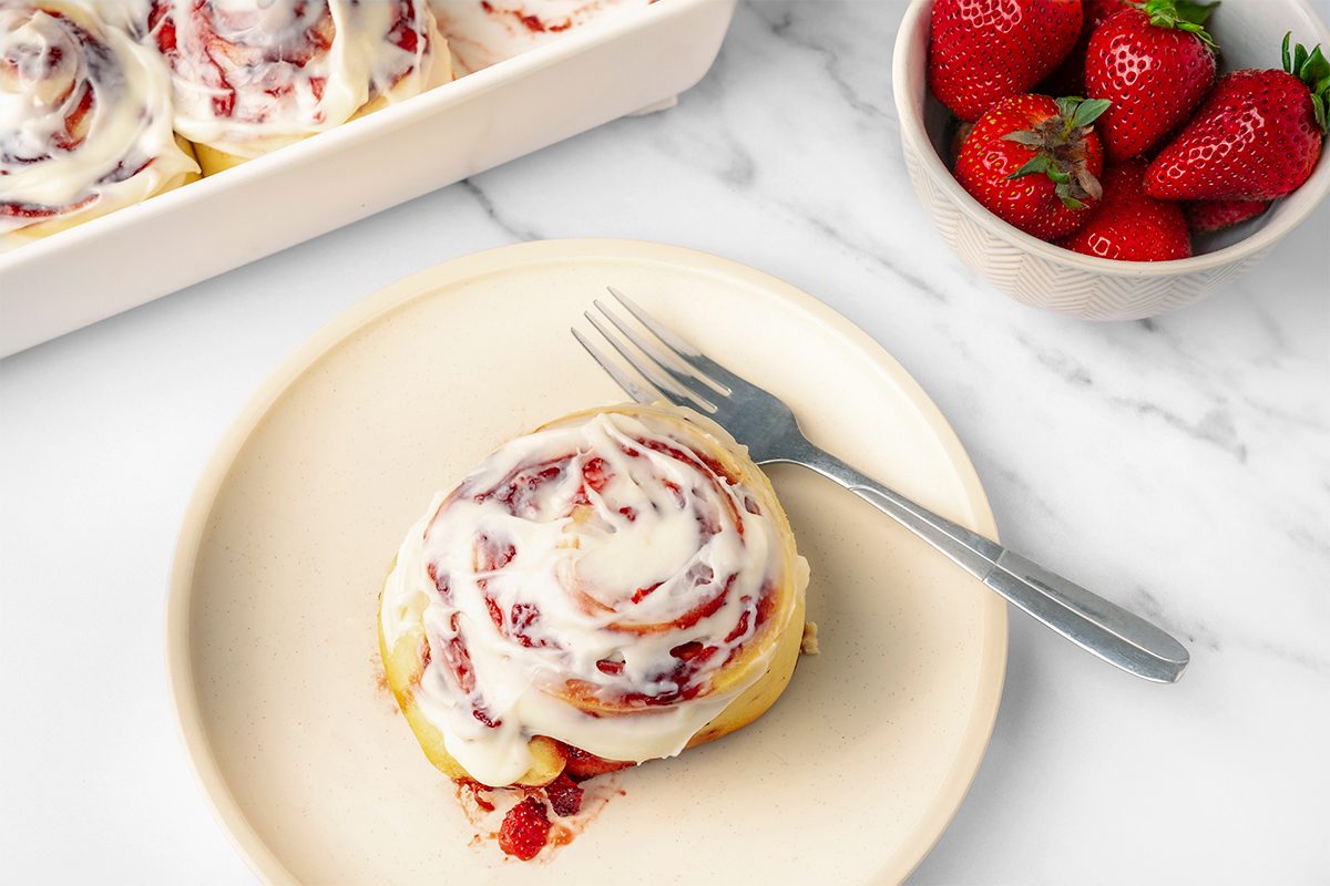 A cinnamon roll with white icing is on a cream-colored plate with a fork beside it. In the background, there are more cinnamon rolls in a baking dish and a bowl of fresh strawberries on a marble surface.