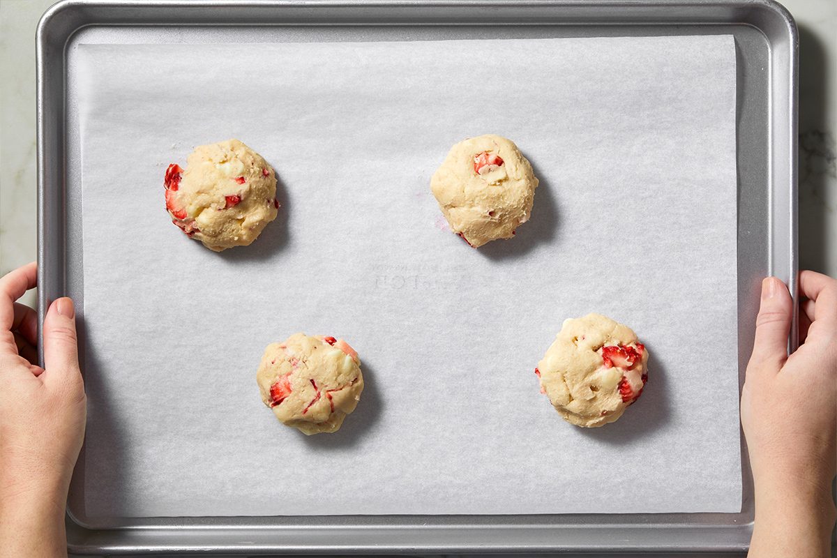 Overhead view of hands holding the prepared strawberry cheesecake cookie dough balls on a parchment paper-lined baking sheet.