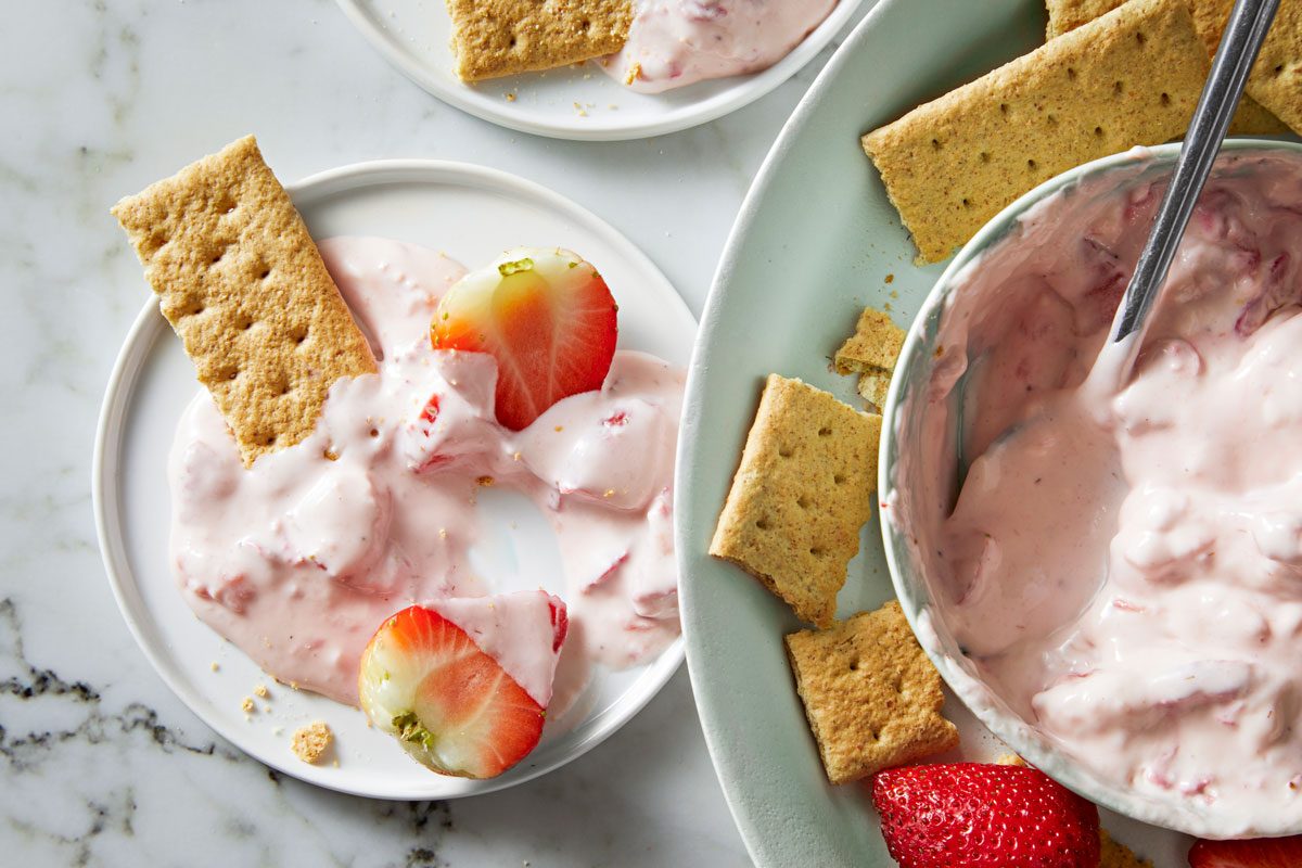 overhead shot of a delicious strawberry dip, served with graham crackers and fresh strawberries; it is served in a white bowl, and it is surrounded by graham crackers and fresh strawberries; the graham crackers are broken into pieces, and the strawberries are sliced;