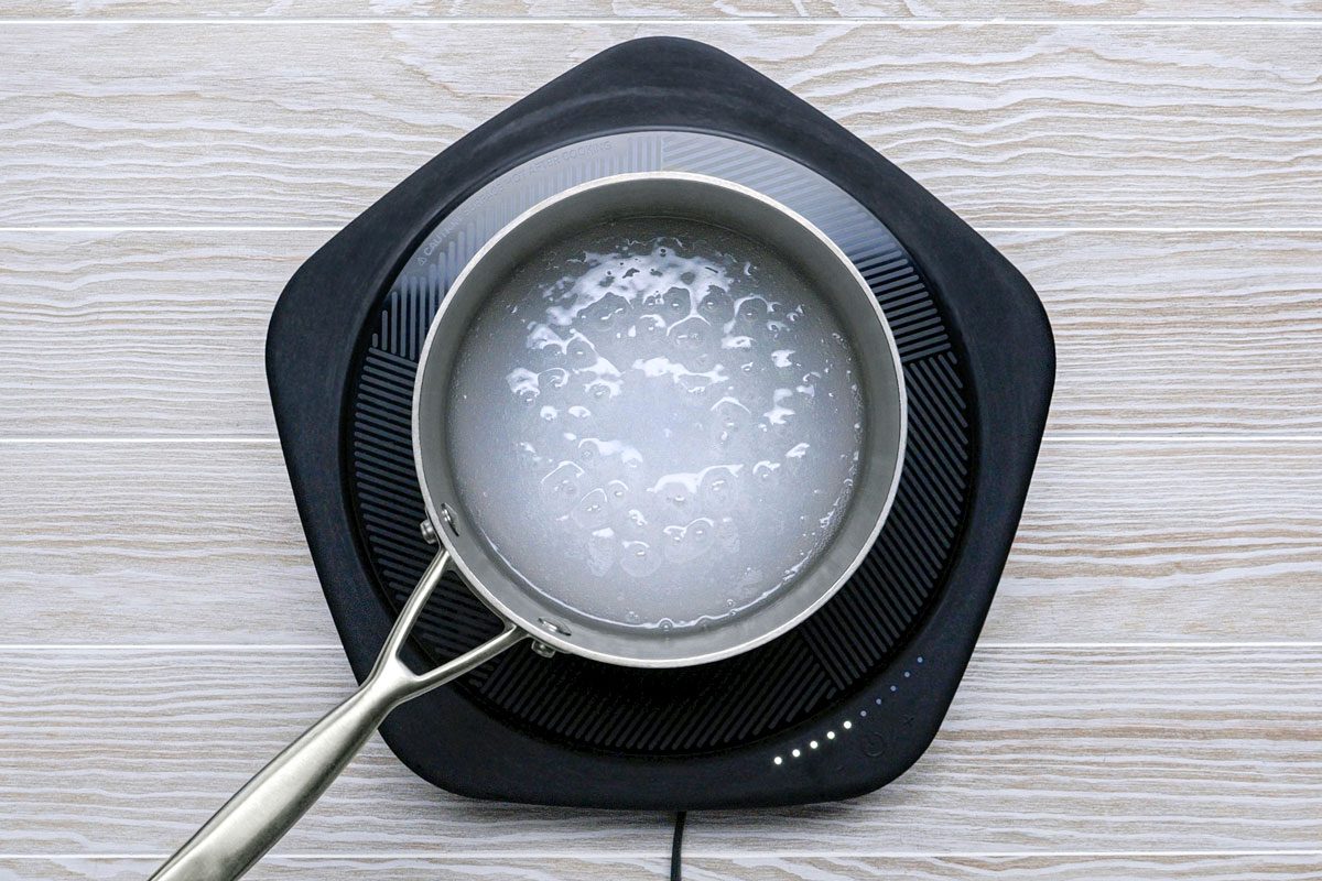 overhead shot of a black electric stovetop with a silver pot sitting on top of it; the pot is filled with water, which is boiling, the stovetop is set on a wooden surface