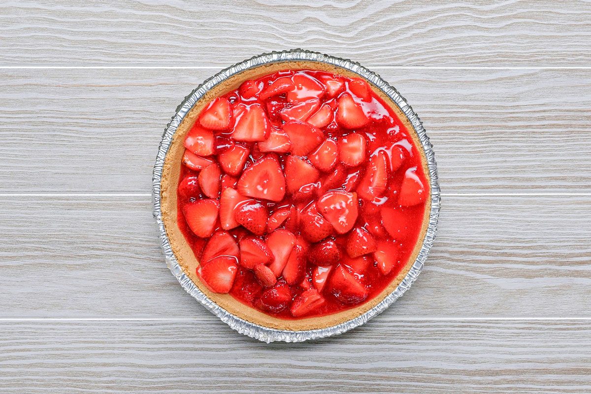 overhead shot of a strawberry pie in a foil pie pan, on a white woodgrain surface; the pie is filled with whole strawberries