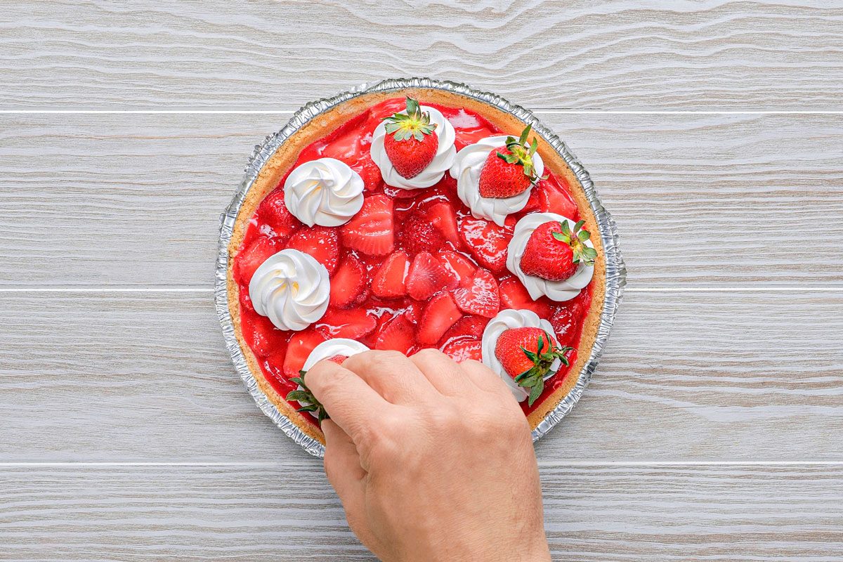 overhead shot of a hand reaches towards a strawberry pie on a white woodgrain table; has a silver foil bottom, a red strawberry filling with whipped cream on top, and strawberries on top of the whipped cream