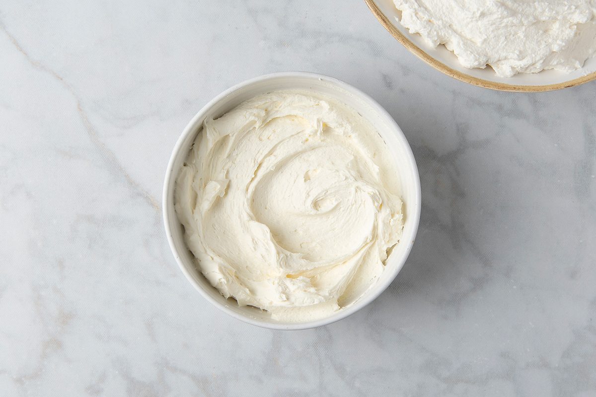 A bowl of creamy white frosting sits on a marble countertop. The frosting is smooth and swirled into a circular pattern. Another bowl with the same frosting is partially visible in the top right corner.