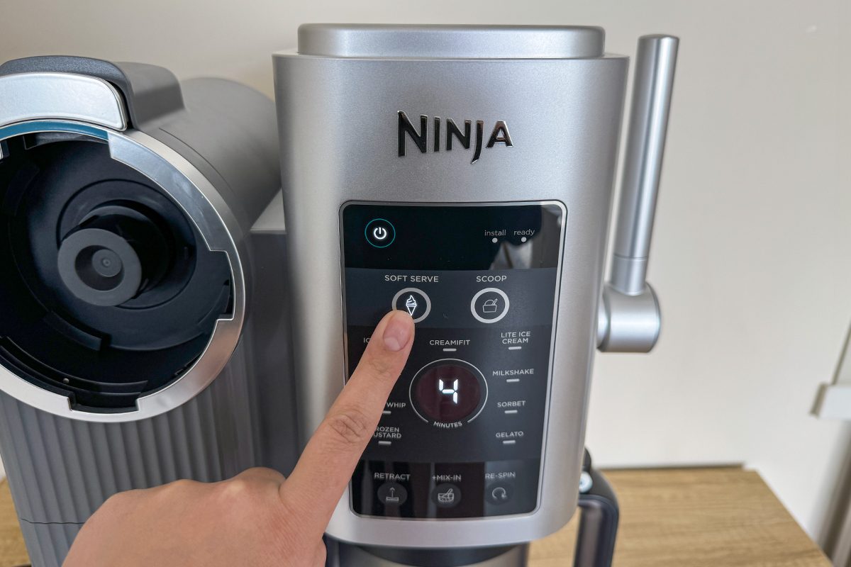 A person pressing the "Espresso" button on a Ninja coffee machine. The control panel displays various options, including cup size and brew settings. The machine is silver and positioned on a wooden surface.