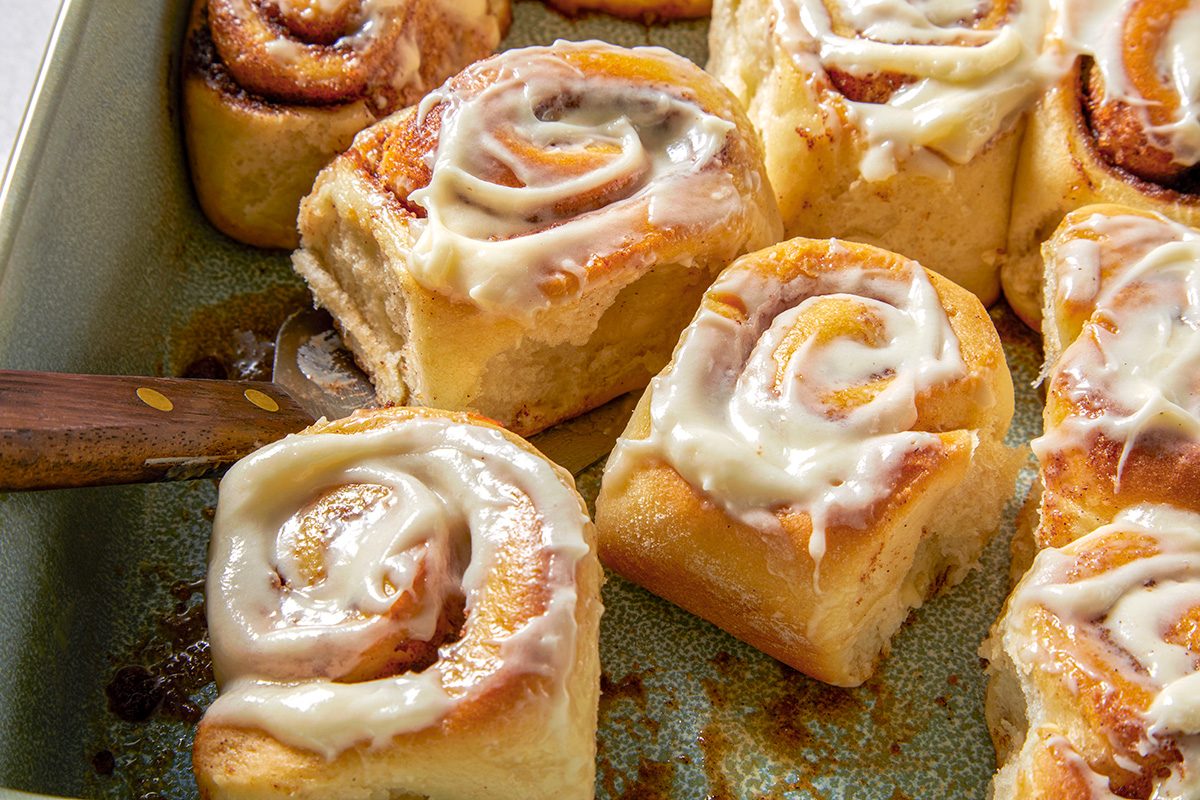 A baking tray filled with cinnamon rolls topped with white icing. A wooden-handled spatula is lifting one roll, revealing its soft, fluffy interior. The rolls are golden-brown and neatly arranged within the tray.