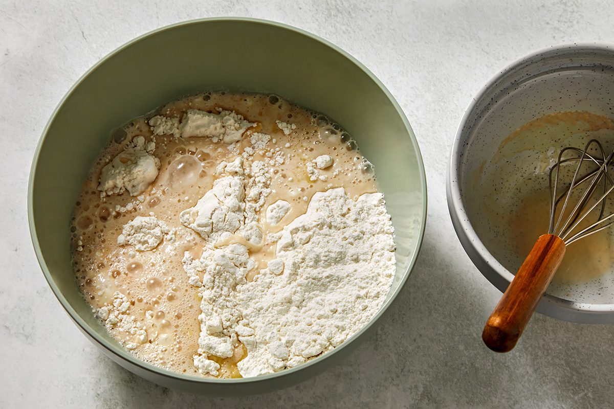 A green bowl contains flour and liquid ingredients being mixed, set on a light surface. Beside it, a gray bowl holds batter and a whisk with a wooden handle.