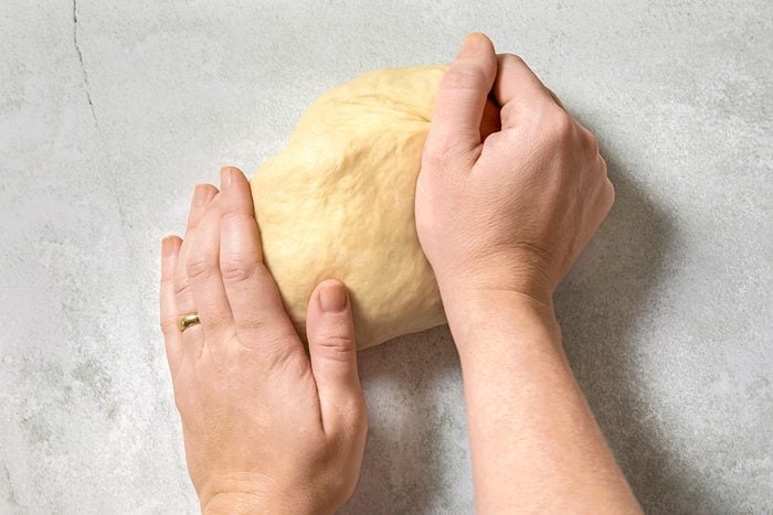 Hands kneading a piece of dough on a light, speckled countertop. One hand holds the dough steady while the other presses into it, showing the kneading process.