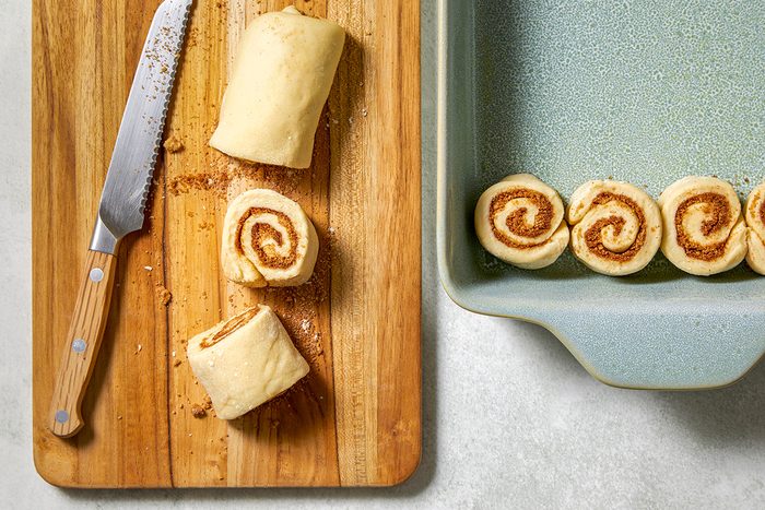 A wooden cutting board with partially sliced cinnamon roll dough next to a knife. Two rolls are visible on the board, and more are neatly arranged in a light green ceramic baking dish on the right.