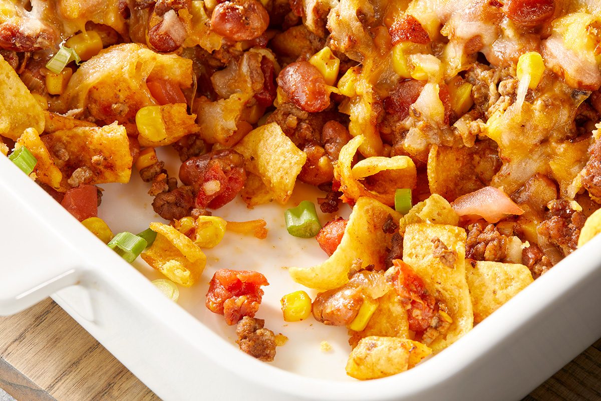 A baked dish filled with corn chips, ground beef, melted cheese, diced tomatoes, corn, and chopped green peppers in a white rectangular casserole dish.