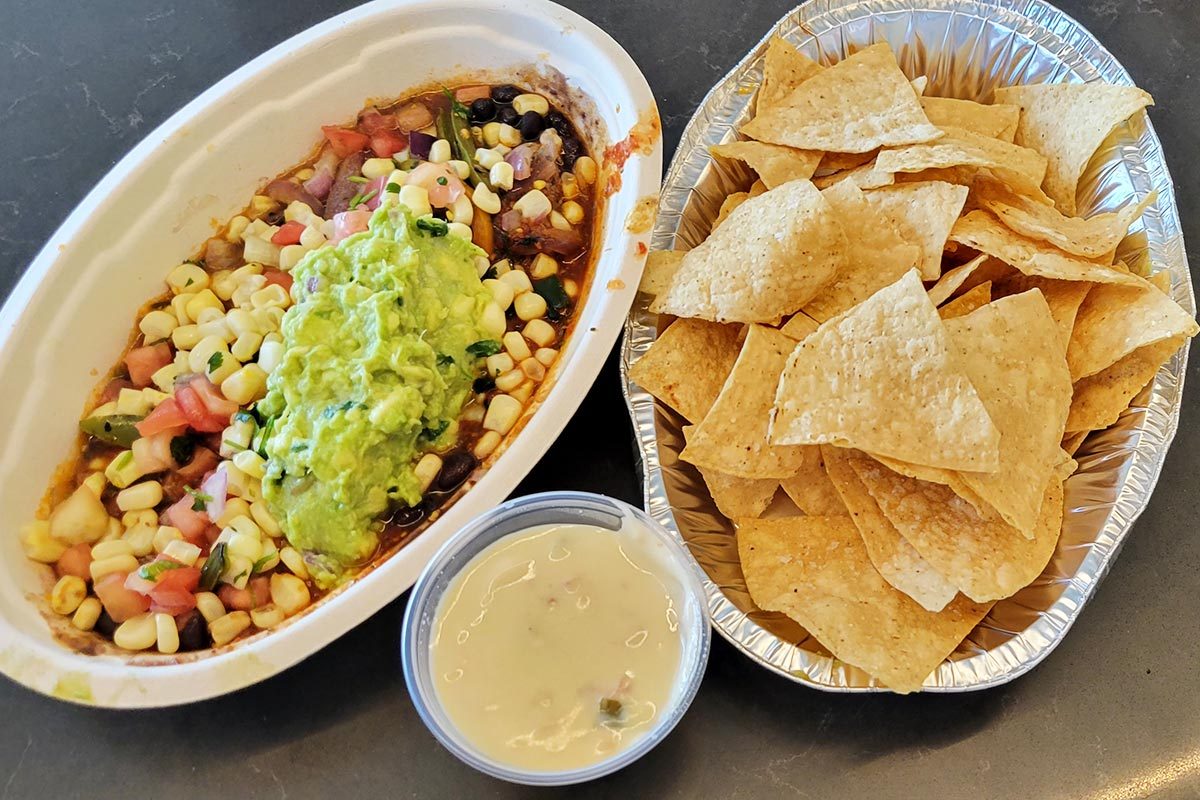 Burrito bowl from Chipotle with guacamole, and a separate cup of queso, next to a lid full of tortilla chips