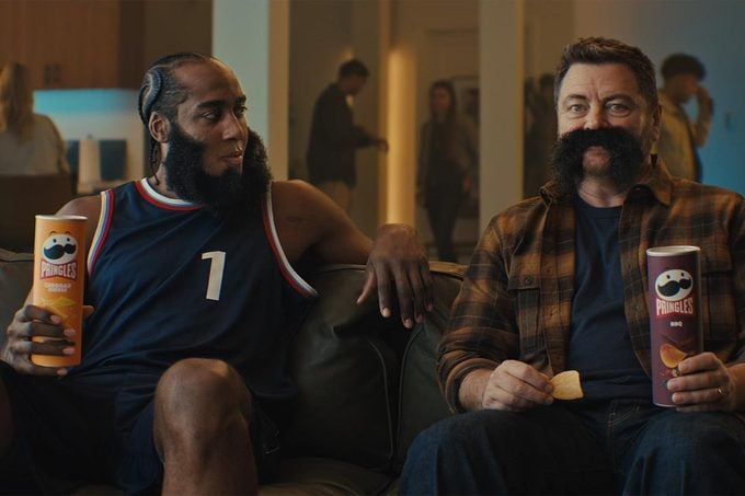 Two men sit on a couch indoors eating pringles