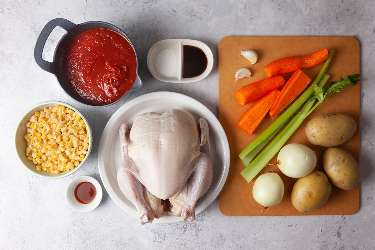 Ingredients for a chicken stew are arranged on a surface: a whole chicken on a plate, a bowl of corn, a bowl of tomato sauce, small dishes with soy sauce and chili, carrots, celery, potatoes, onions, and garlic cloves.