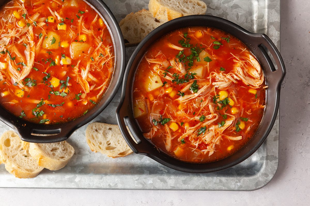 Two black bowls of chicken vegetable soup with shredded chicken, corn, and herbs. The bowls are on a metal tray with slices of crusty bread on the side. The soup appears colorful and hearty.