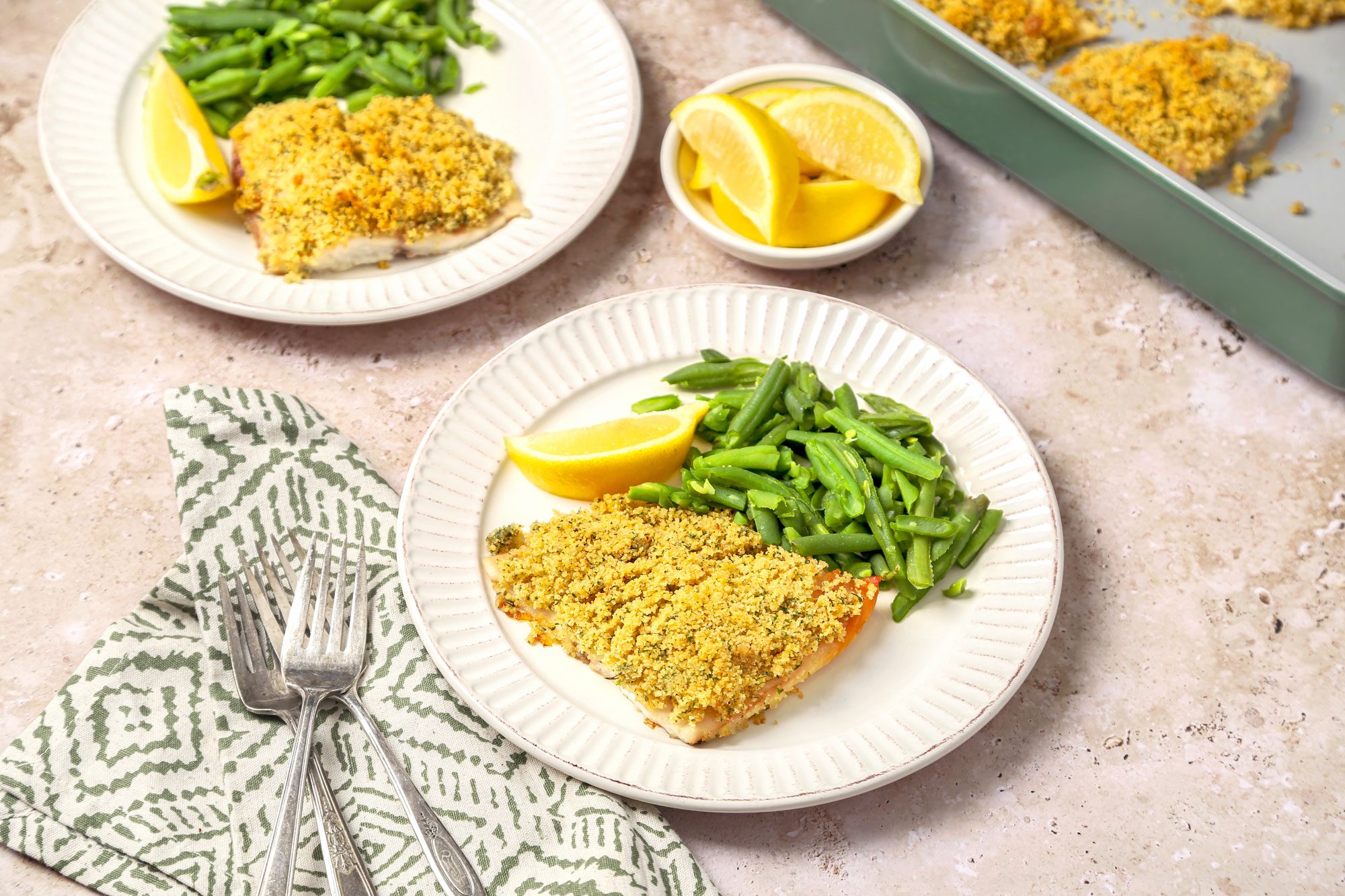 3/4th shot of Baked lemon Haddock shot on a beige marble surface; Baked fish served on plates with lemon wedge and green beans; two silver forks on light green pattern napkin