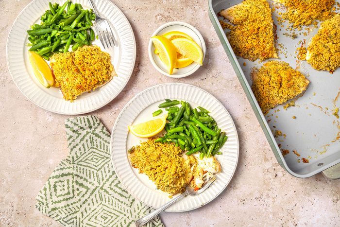 overhead shot of Baked lemon Haddock shot on a beige marble surface; Baked fish served on plates with lemon wedge and green beans; two silver forks on each plate with light green pattern napkin