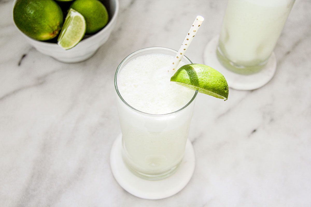 A frosty glass filled with Brazilian Lemonade, garnished with a lime wedge and a dotted straw, sits on a marble countertop.