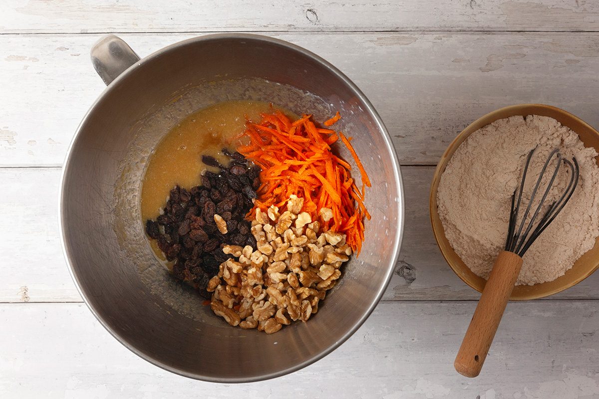 A mixing bowl with grated carrots, raisins, and chopped nuts on top of a batter. A separate bowl with flour and a whisk is beside it on a white wooden surface.