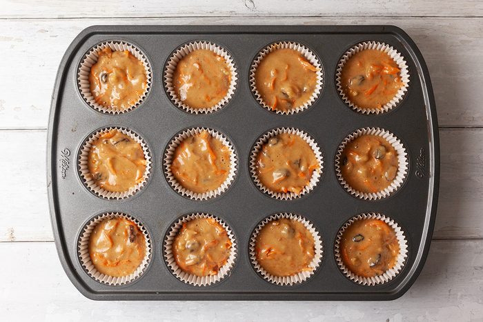 Muffin tray containing twelve muffin liners filled with brown batter mixed with chunks, placed on a light wooden surface.