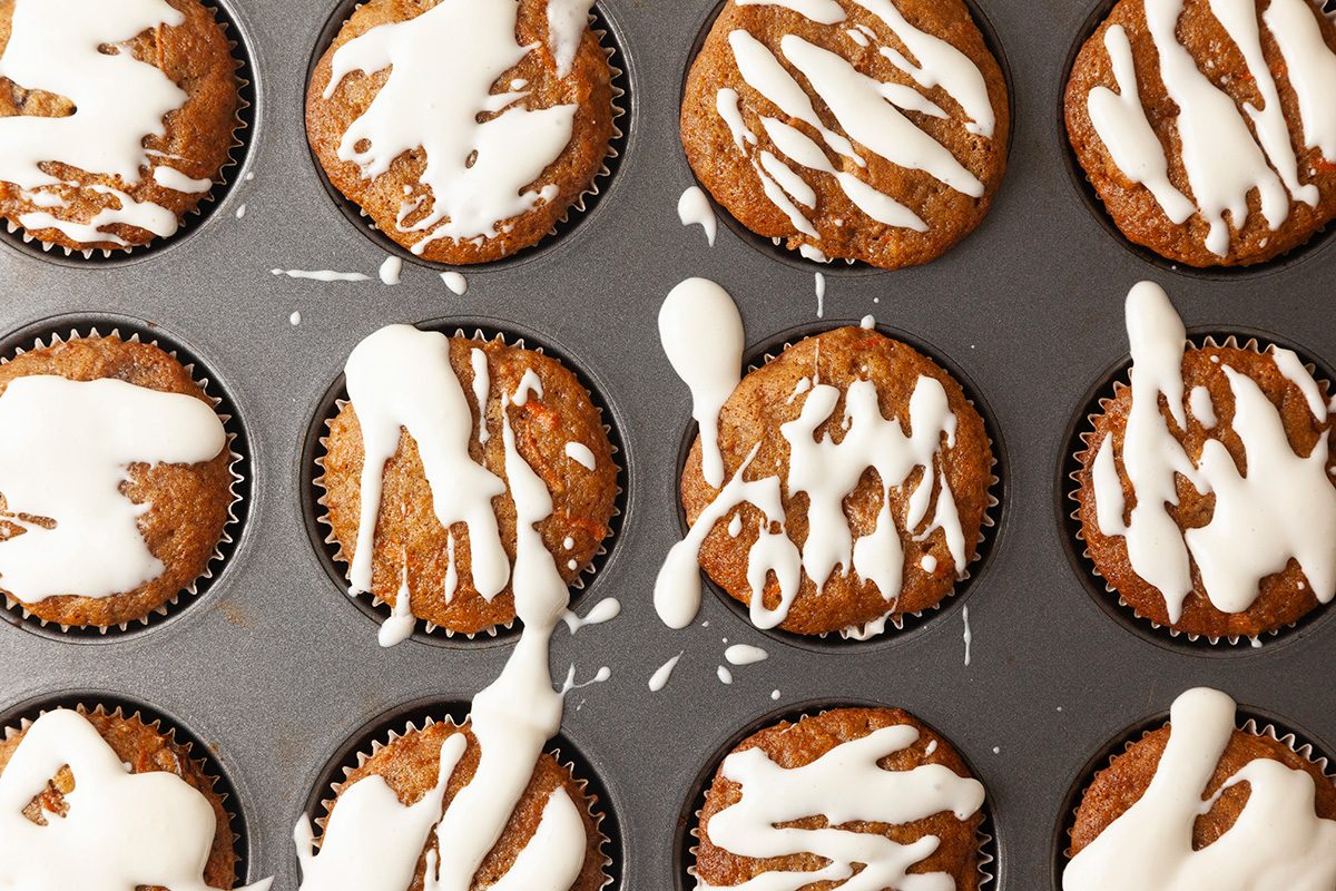 A muffin tray filled with baked muffins drizzled with white icing. The muffins are golden brown and the icing is randomly spread across the tops, creating a homemade appearance.