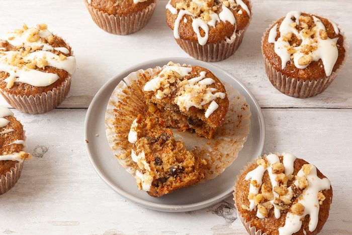 Seven muffins topped with white icing and nuts are on a white wooden surface. One muffin is cut in half on a gray plate, revealing a moist interior with visible nut pieces. The muffins have a light golden-brown color.