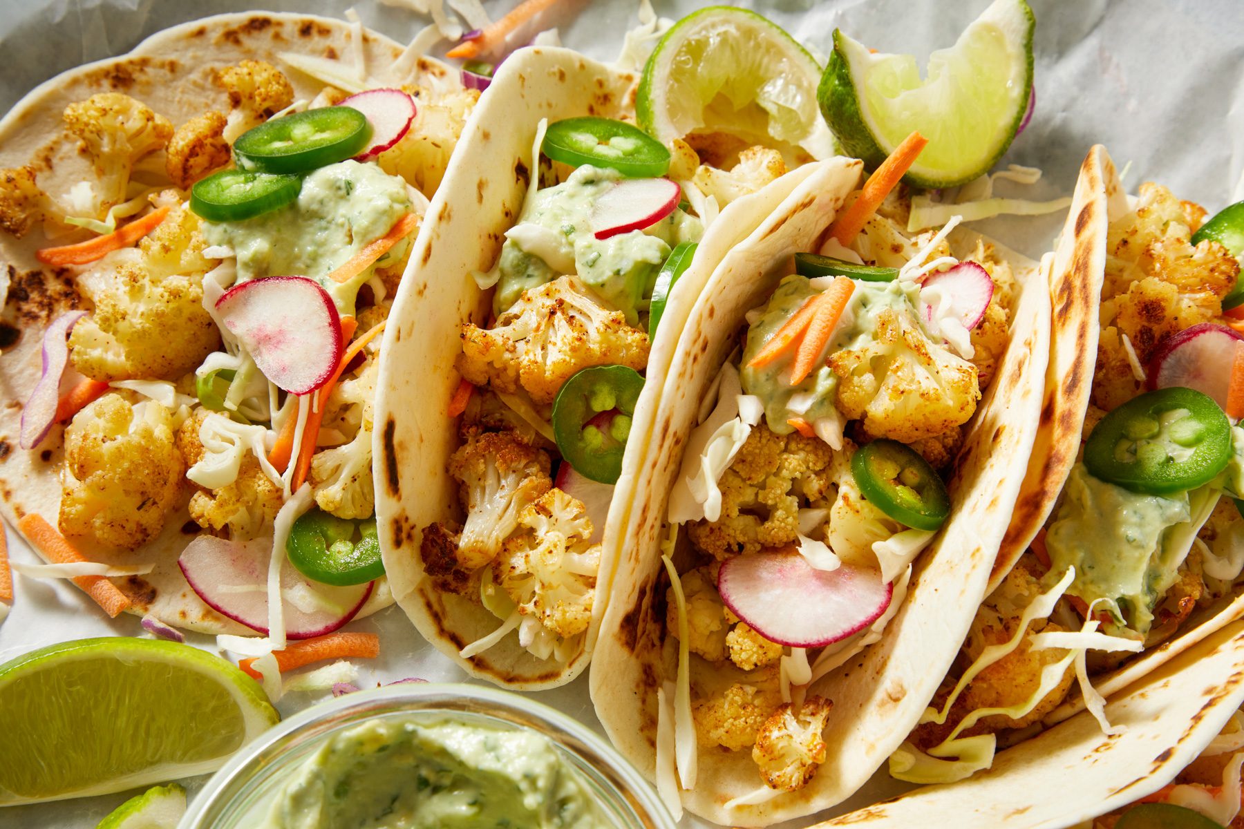 Close-up shot of Cauliflower Tacos; Serve in tortillas with coleslaw mix avocado crema and toppings; lime wedges; dark blue surface;