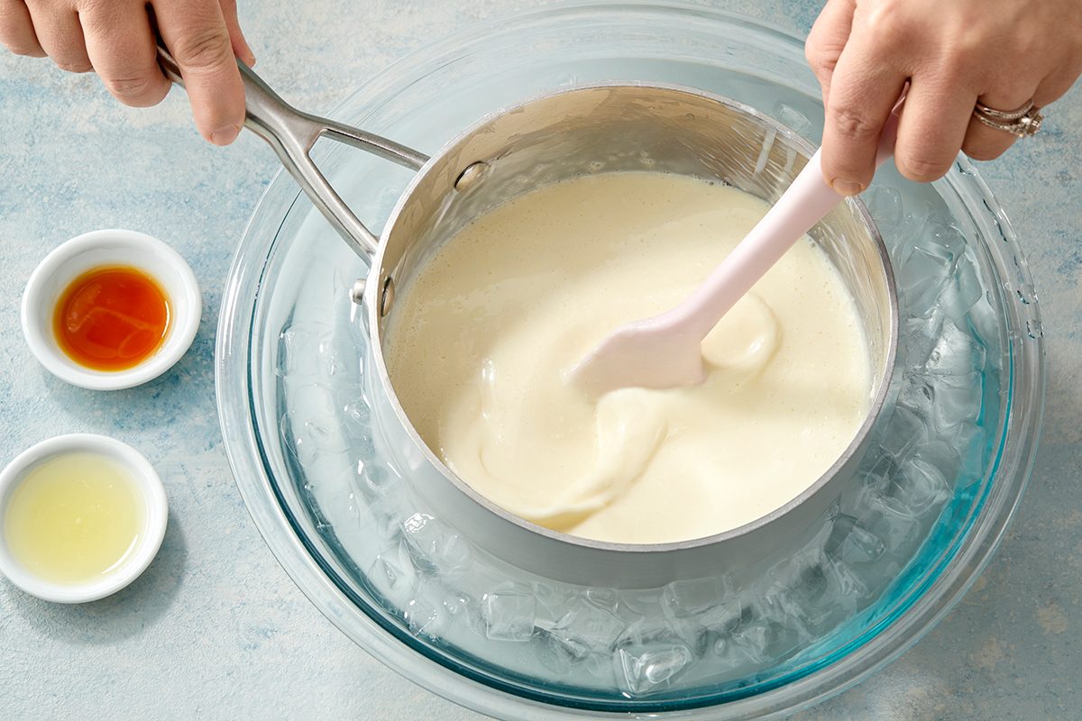 Hands holding a saucepan with white chocolate ganache over a bowl of ice. A spatula stirs the ganache. Small bowls of vanilla extract and lemon juice are nearby on a light blue surface.