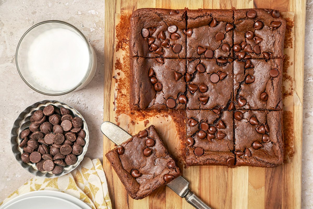 A batch of Chickpea chocolate brownies on a wooden board, cut into squares. One piece is on a knife. 