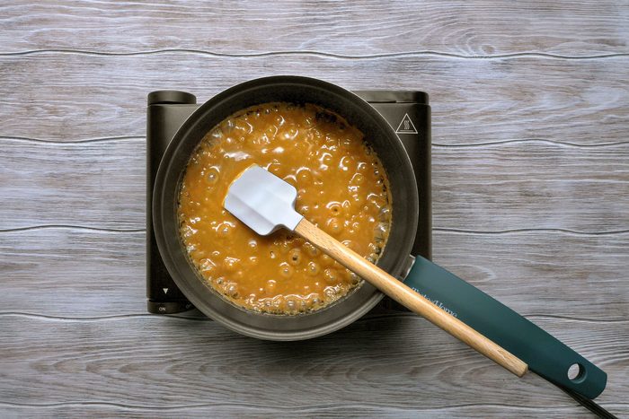 overhead shot of a pot of caramel being cooked on a portable electric stovetop, likely for making a dessert; The pot is filled with golden-brown caramel, bubbling gently as it simmers; A white silicone spatula is resting in the caramel, with a wooden handle that extends out of the pot
